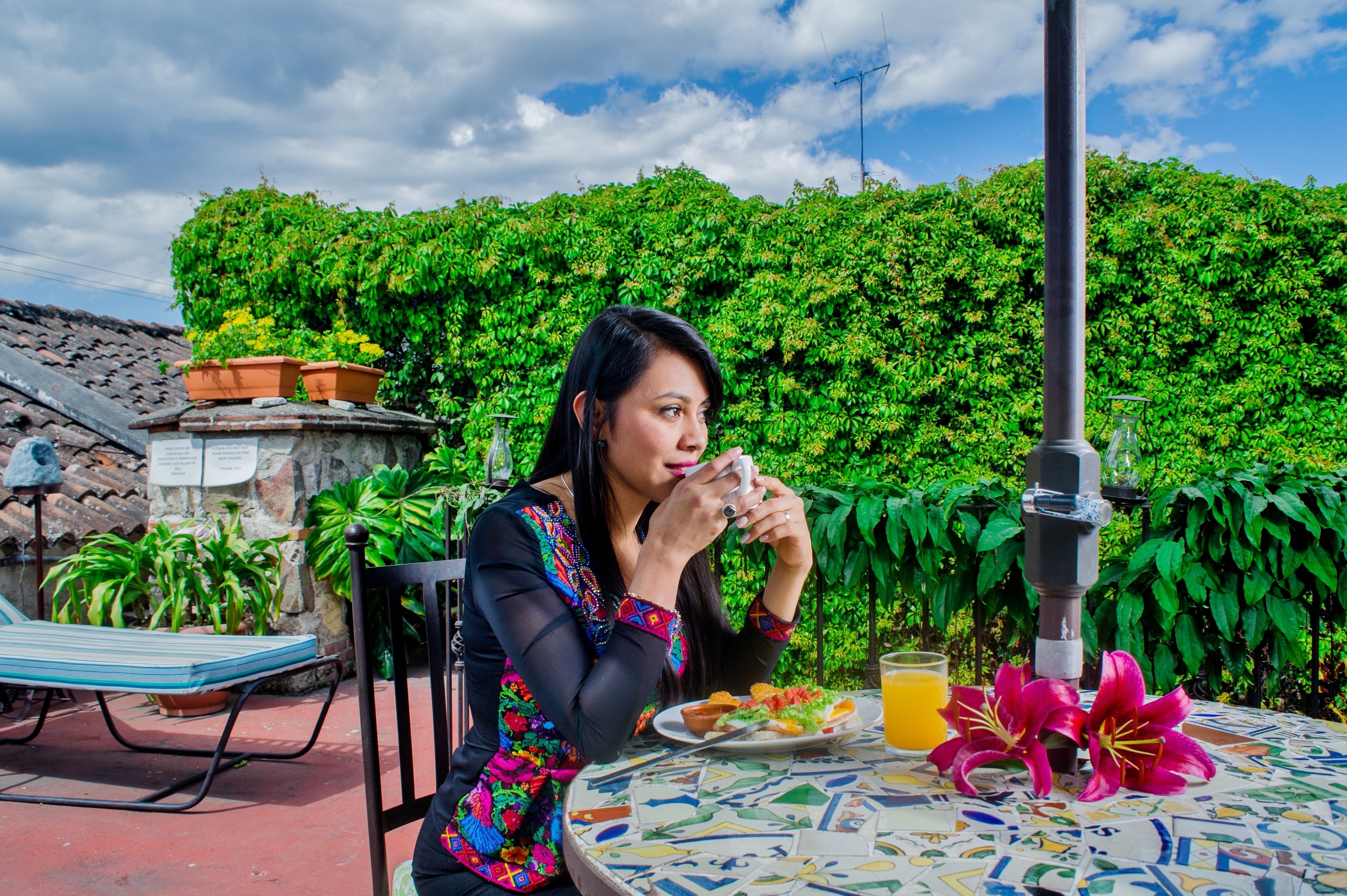 A person sitting at a table with a plate of food and a glass of orange juice, outdoors with a green hedge and tiled roof in the background.