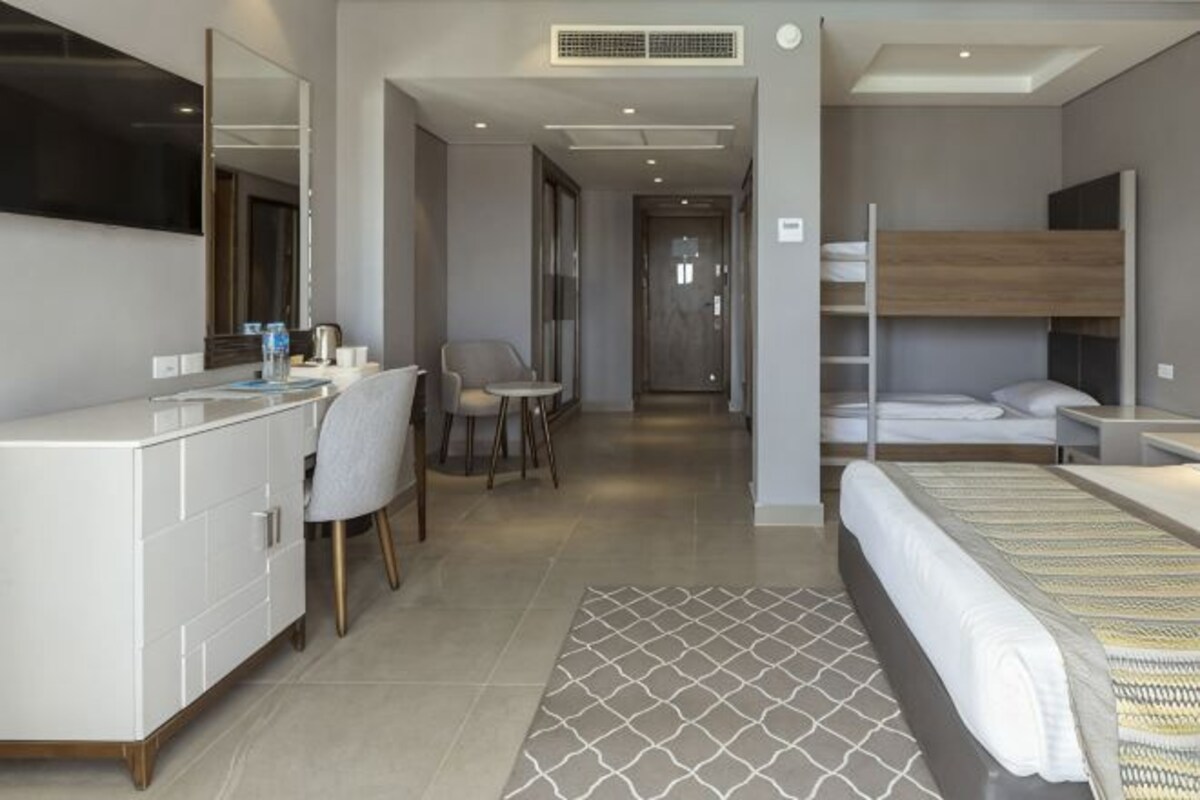A modern hotel room with a large bed, a bedside table, a chair, a desk, a vanity with a mirror, and a television.
