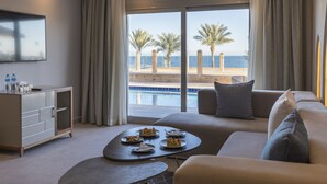 Ein modernes Hotelzimmer mit einer Sitzecke, einem Couchtisch mit Snacks, einem Fernseher und Blick auf einen Pool und Palmen.