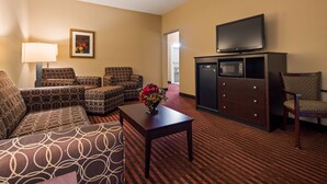 A hotel room with a sofa, armchair, and a small table with a vase of flowers. There is a flat-screen TV mounted on the wall and a minibar. - Best Western Plus Louisa (Louisa)