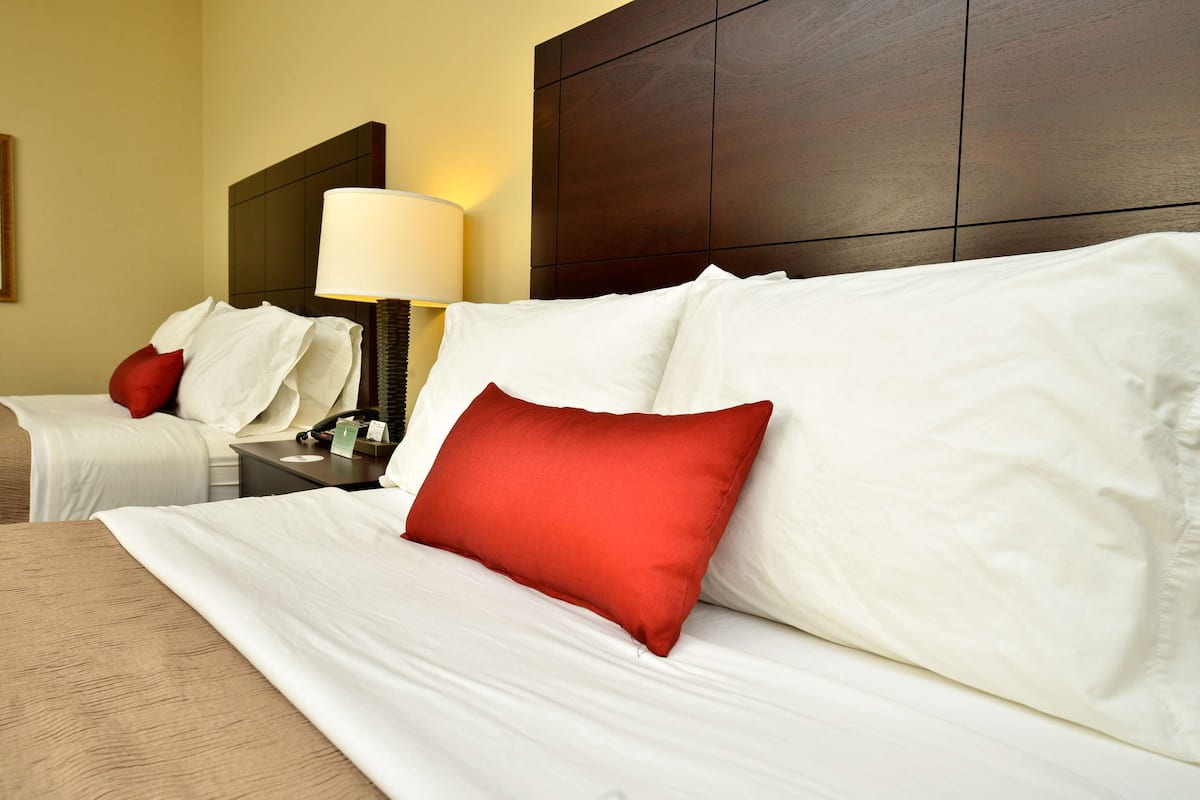 Quarto de hotel com duas camas, cada uma com roupa de cama branca e almofadas vermelhas. Há uma mesa de cabeceira com uma luminária entre as camas.