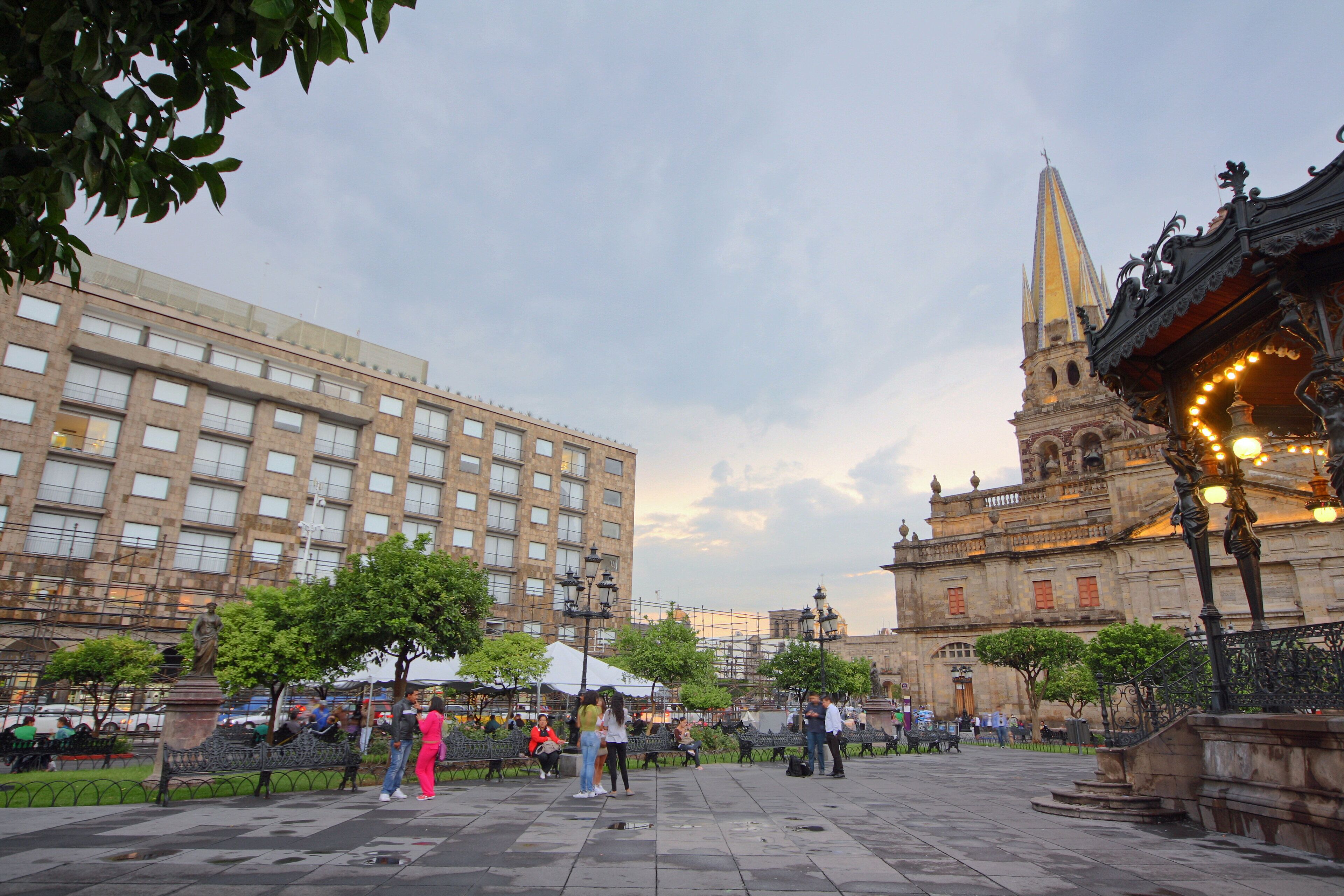 A public square with benches, a statue, and a historic building with a tall spire.