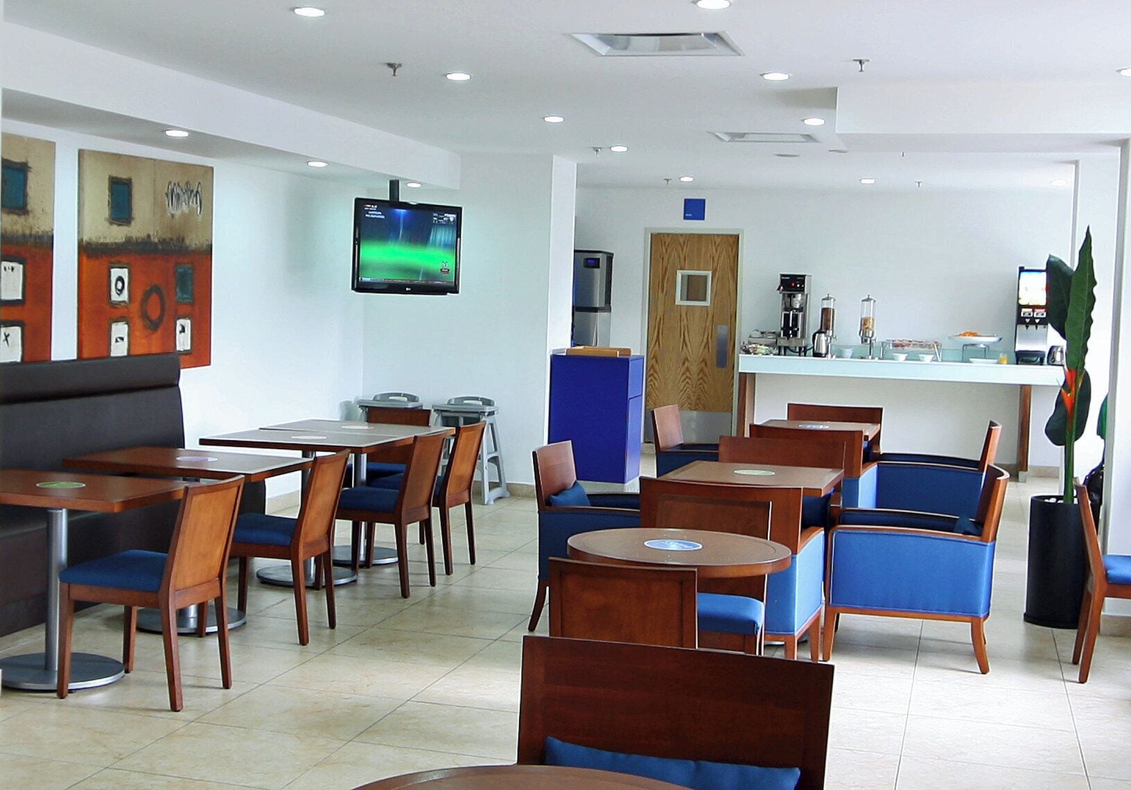 Un restaurante moderno con mesas de madera y sillas azules, un televisor en la pared y una zona de cocina al fondo.