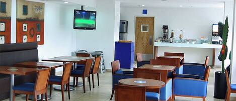 Un restaurante moderno con mesas de madera y sillas azules, un televisor en la pared y una zona de cocina al fondo.