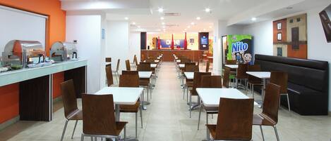 A cafeteria with multiple tables and chairs, a counter with food preparation equipment, and a vending machine.