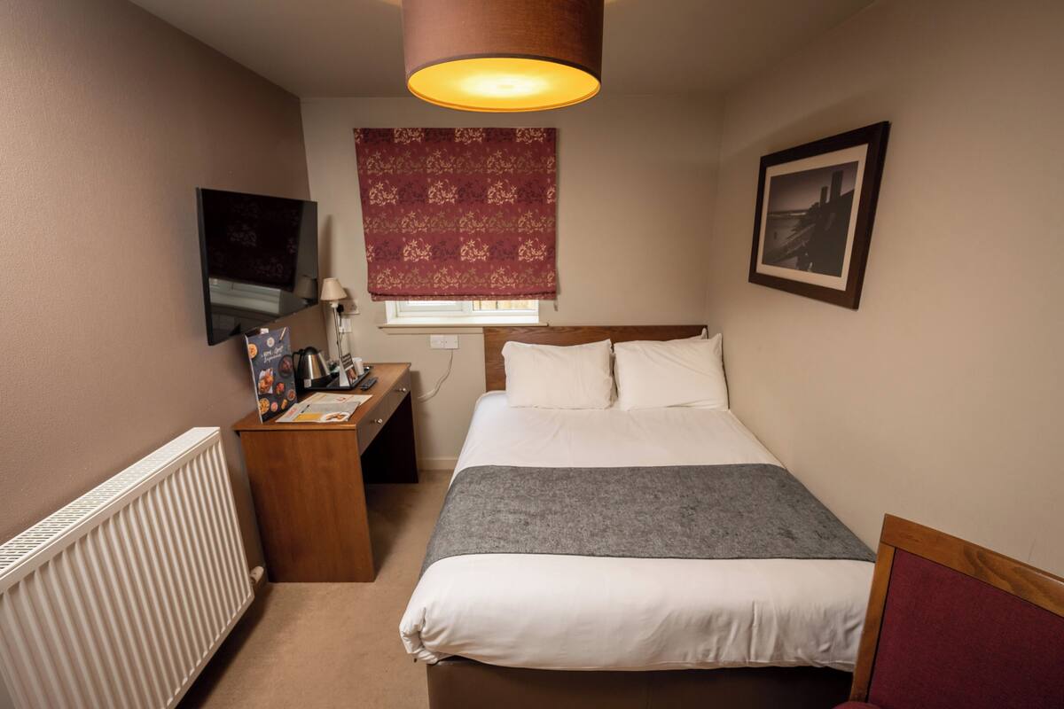 A bedroom with a bed, a desk, a lamp, a radiator, a window with a curtain, and a picture on the wall.