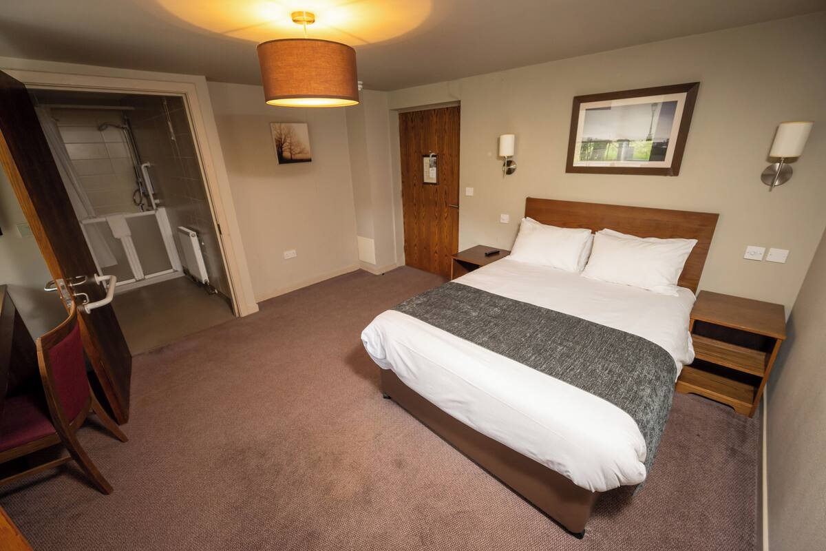 A hotel room with a large bed, a wooden headboard, a standing lamp, a framed picture on the wall, and a door leading to a bathroom.