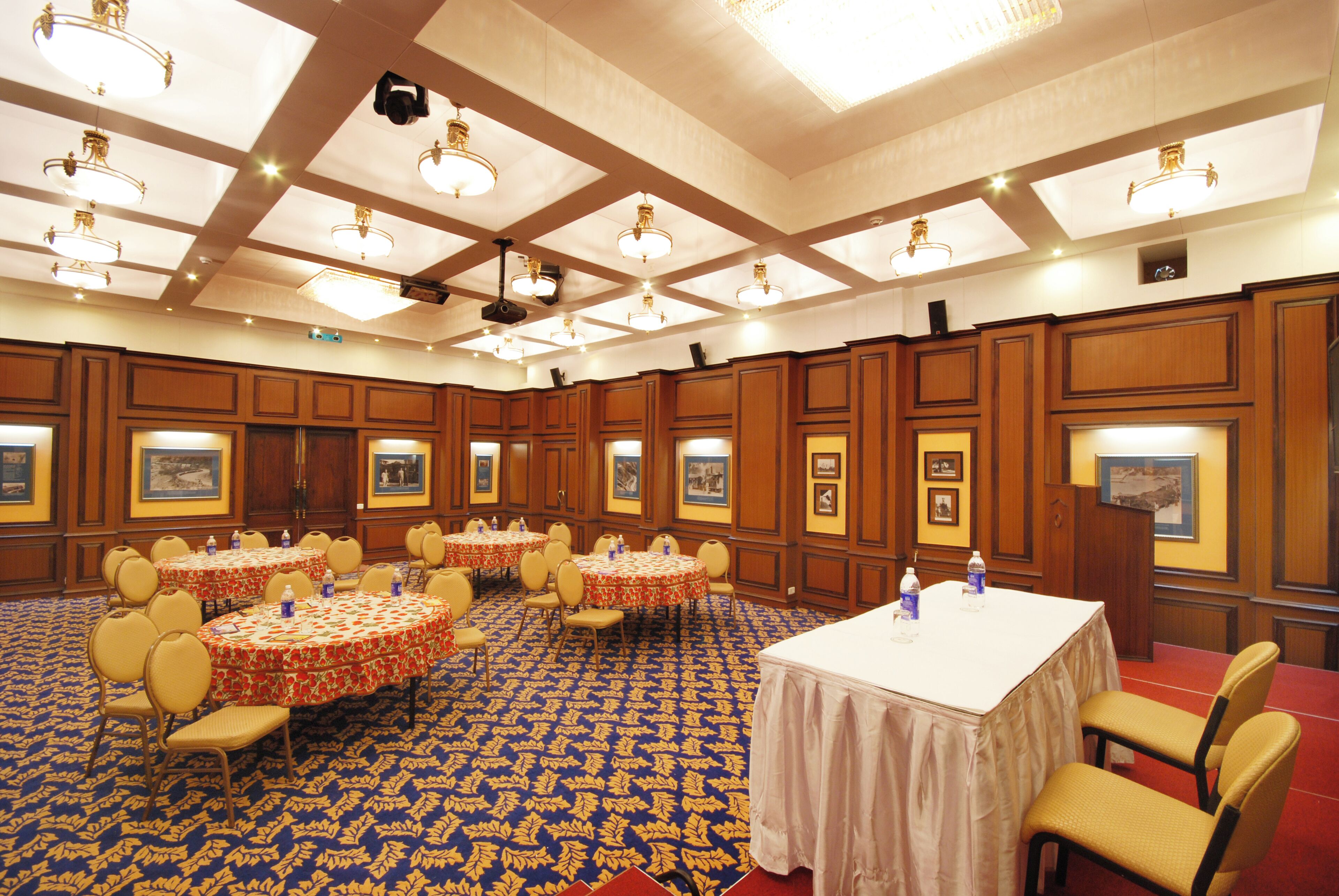 A large conference room with multiple round tables and chairs, a patterned carpet, and wooden paneling on the walls.