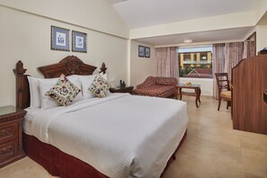 A hotel room with a large bed, a wooden nightstand, a wooden wardrobe, a patterned sofa, a wooden chair, and a window with curtains. - MAYFAIR Heritage, Puri (Puri)