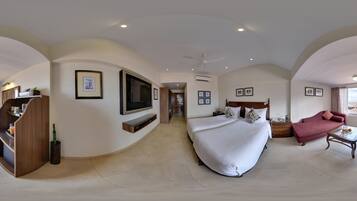 Quarto espaçoso com cama grande, TV na parede, área de estar com sofá e poltrona, e mesa de jantar.