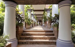 Un paseo cubierto con escalones de piedra y columnas blancas, rodeado de vegetación.