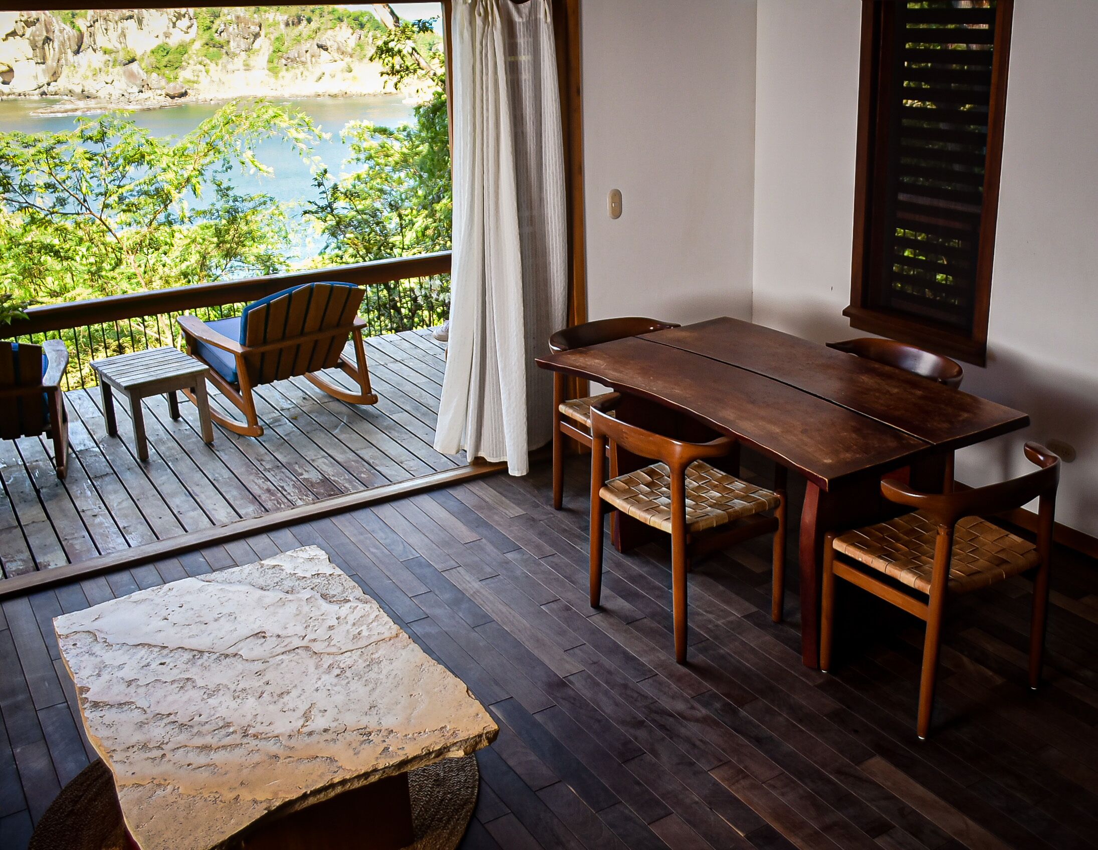 Terraza de madera con una mesa, sillas y una mecedora, con vista a un lago.
