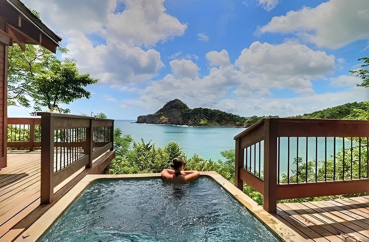 A wooden deck with a hot tub overlooking a clear blue sea and a distant island.
