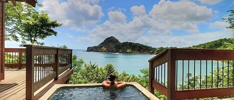 A wooden deck with a hot tub overlooking a clear blue sea and a distant island.
