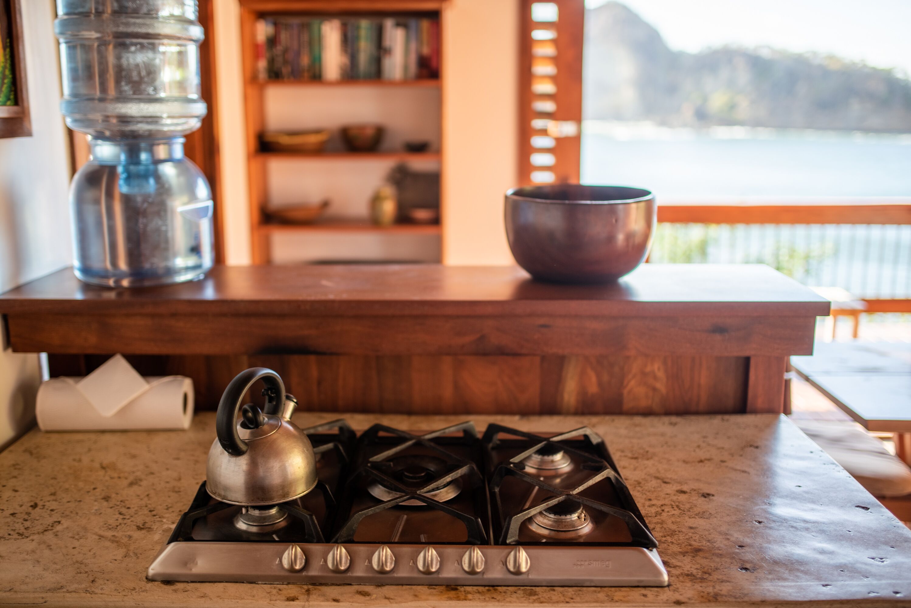Una cocina con estufa a gas, hervidor, olla de metal y repisa de madera con libros y un tazón.