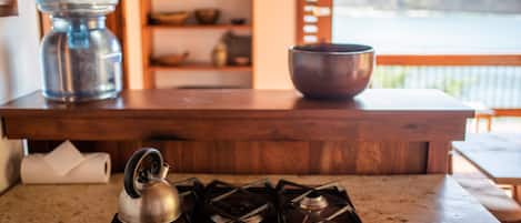 Una cocina con estufa a gas, hervidor, olla de metal y repisa de madera con libros y un tazón.
