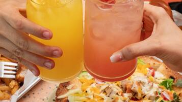 A person holding two drinks, one orange and one pink, with a salad and a piece of bread on the table.