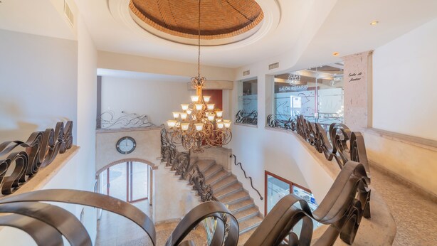Uma escadaria imponente com um lustre central, que leva a um segundo andar com um salão e vista para o exterior.