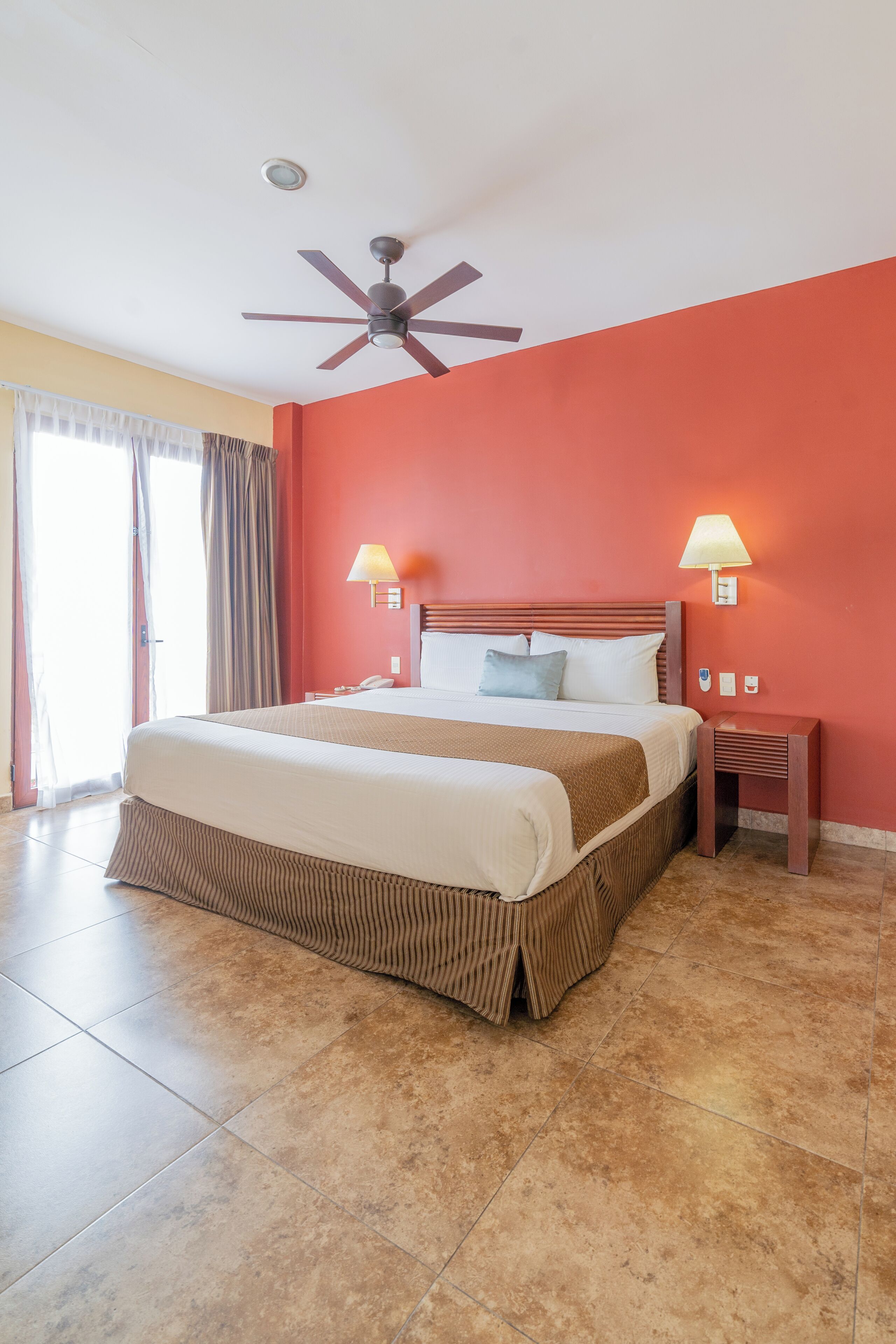 Une chambre d’hôtel avec un grand lit, deux lampes de chevet, un ventilateur de plafond et une vue sur l’extérieur par une fenêtre munie de rideaux.