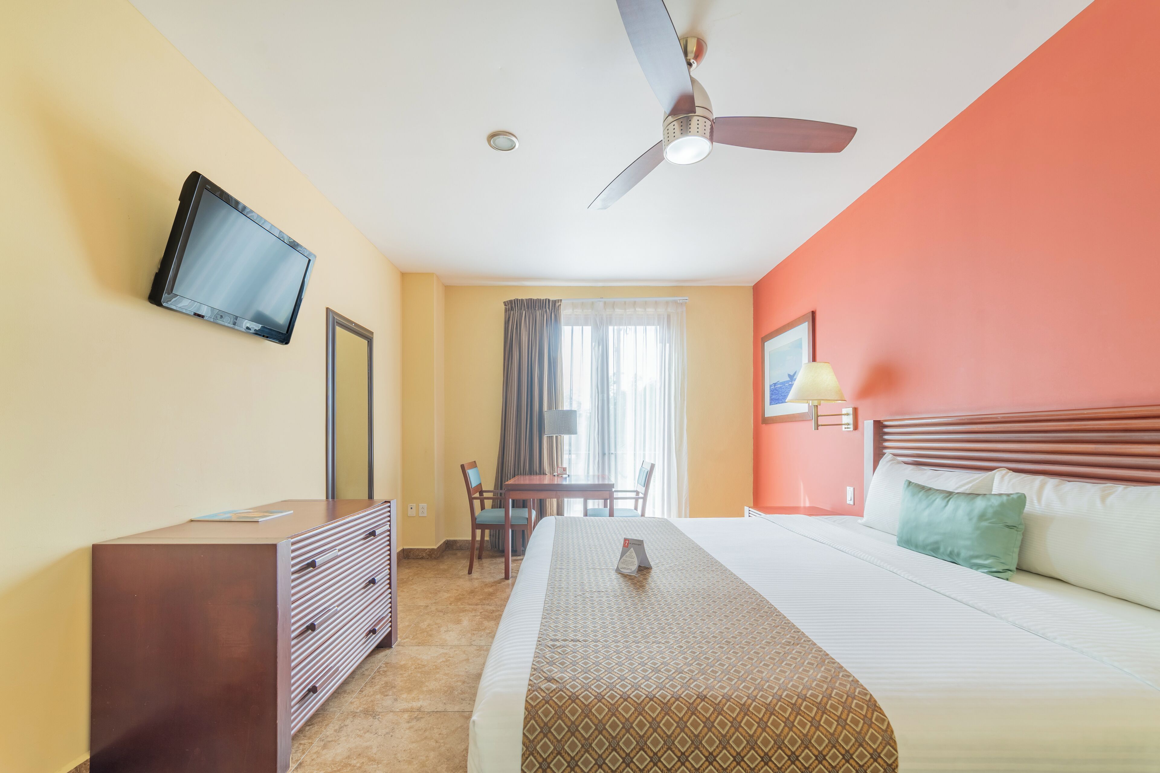 Habitación de hotel con cama, escritorio, silla, televisor montado en la pared y ventilador de techo.