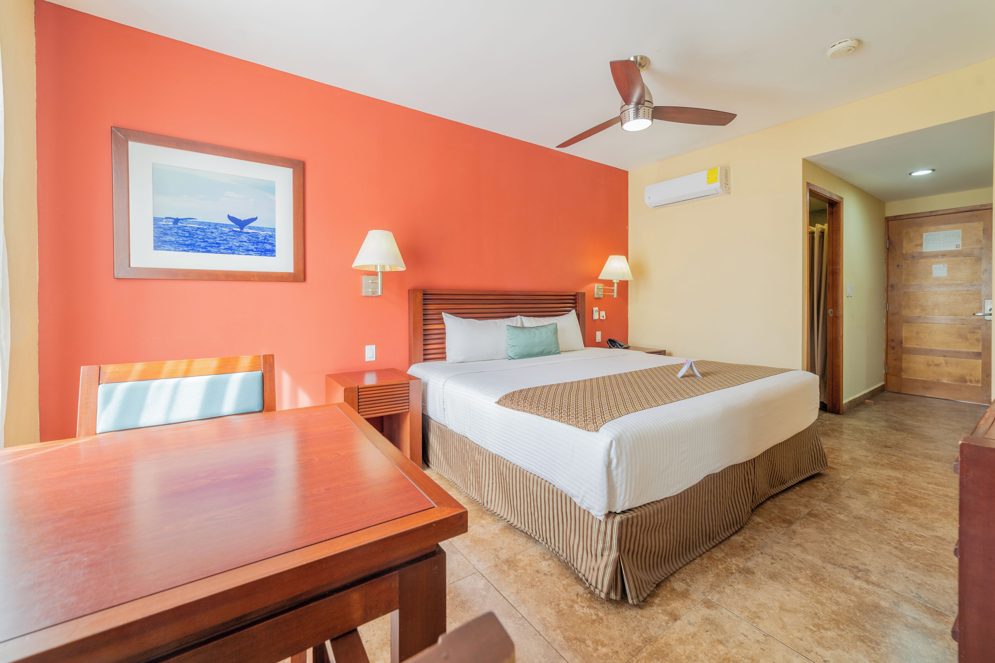 Une chambre d’hôtel avec un lit, des lampes de chevet, un ventilateur de plafond, une table en bois et un tableau encadré au mur.
