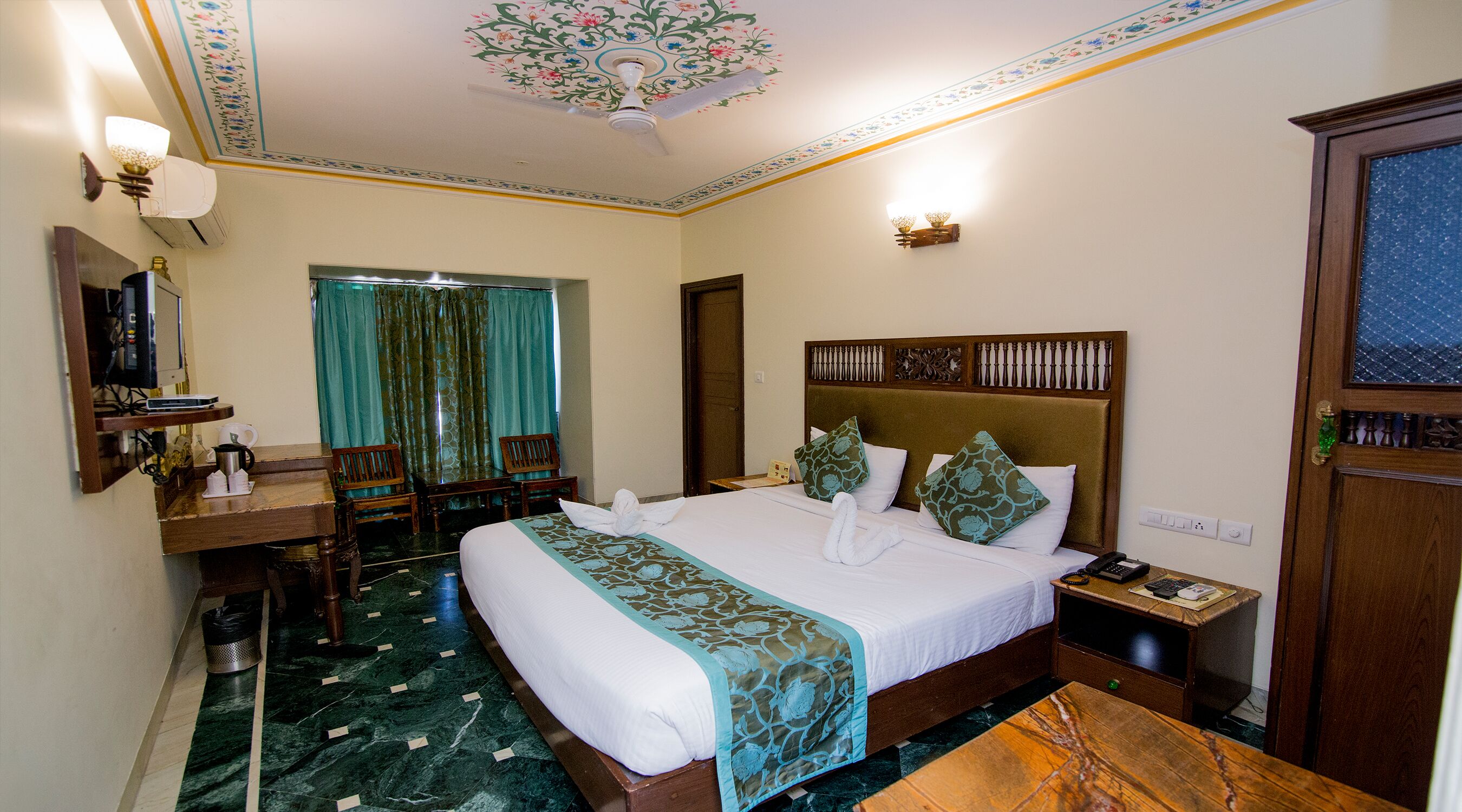 A hotel room with a large bed, a desk with a chair, a telephone, and a decorative ceiling.