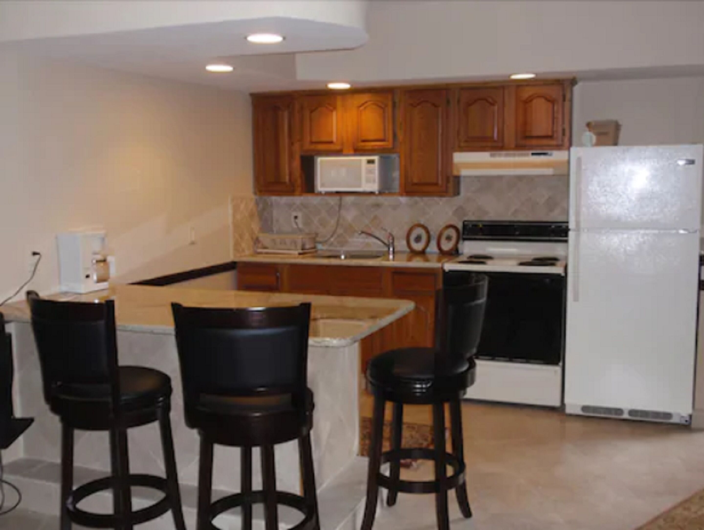A kitchen with wooden cabinets, a white refrigerator, a microwave, and a stove. There are three black bar stools.