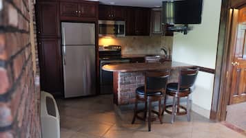 Una cocina con armarios de madera oscura, un refrigerador de acero inoxidable, un microondas, un horno y un televisor integrado. Hay dos taburetes de bar y una pared de acento de ladrillo.