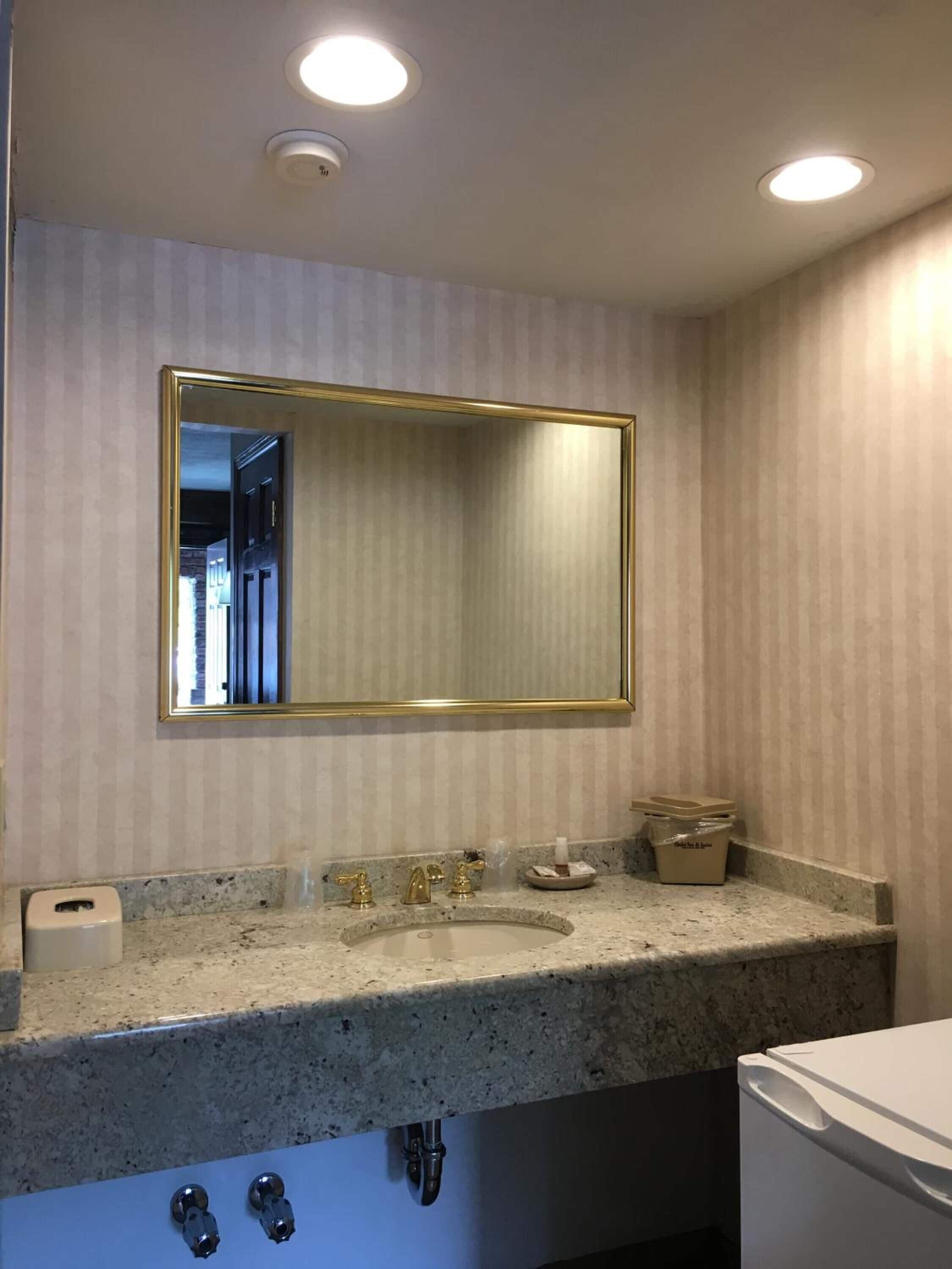 A bathroom with a large mirror, granite countertop, and a sink with gold fixtures.