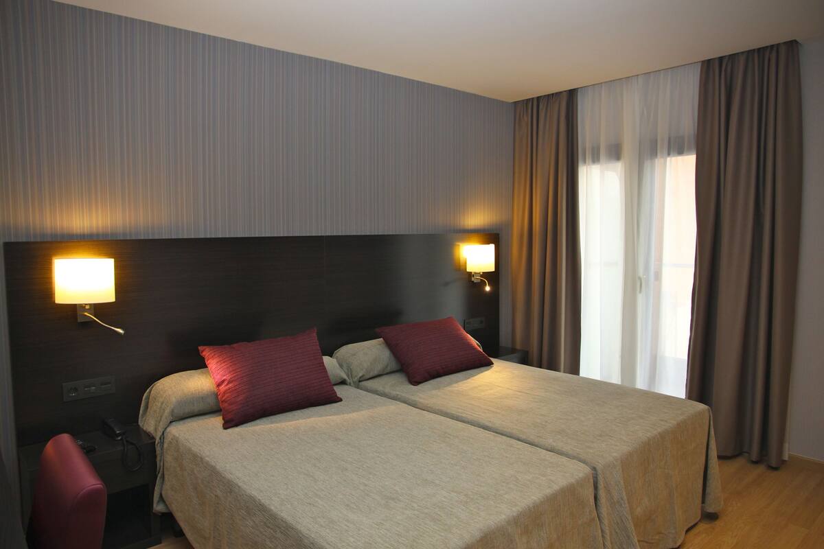 A double bed with two pillows, a bedside table with a lamp, and a window with curtains.