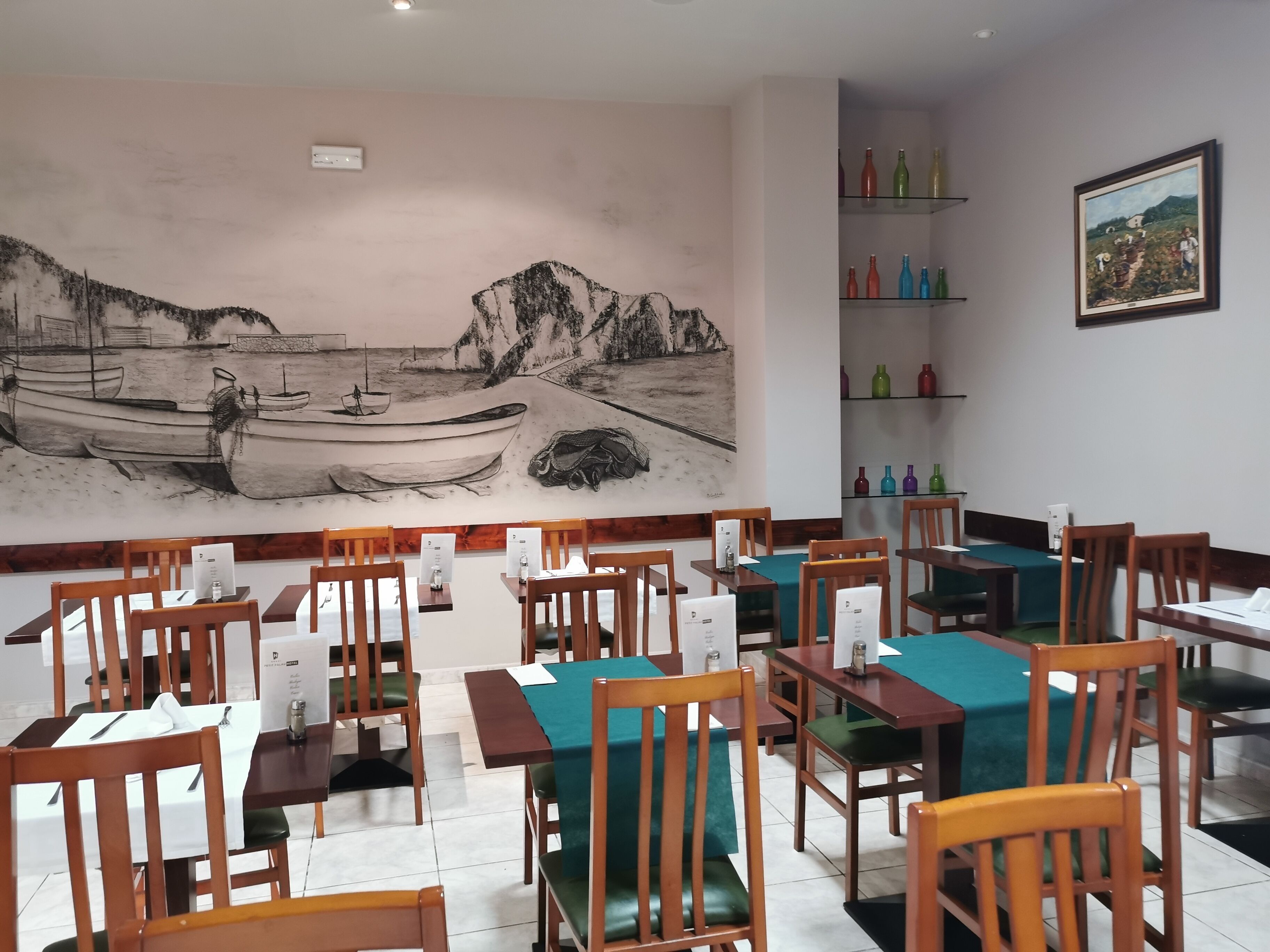Un restaurante con sillas y mesas de madera, un mural de barcos en la playa y estantes con botellas de colores.