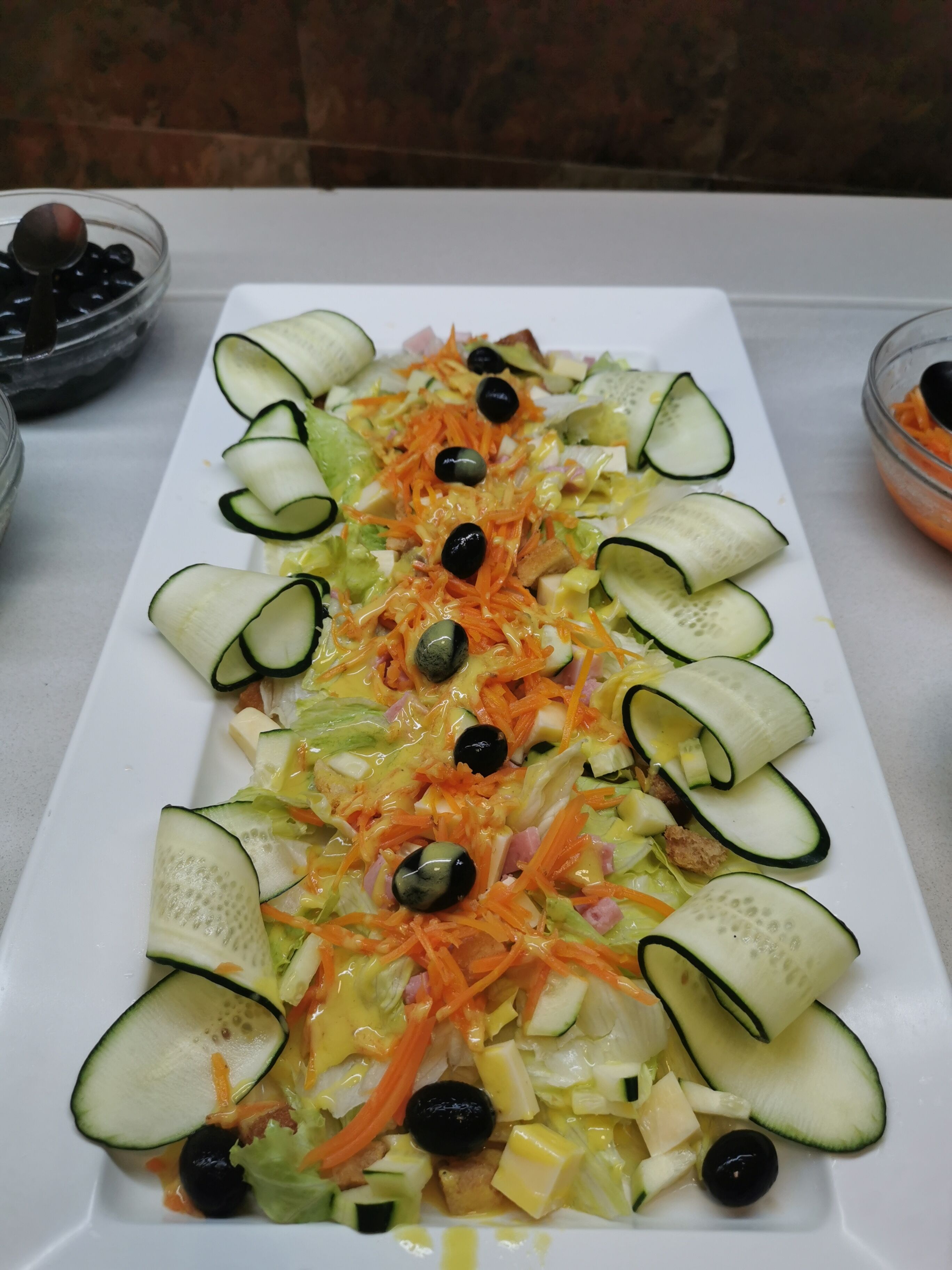 A rectangular white plate with a salad consisting of cucumbers, carrots, olives, and other vegetables.