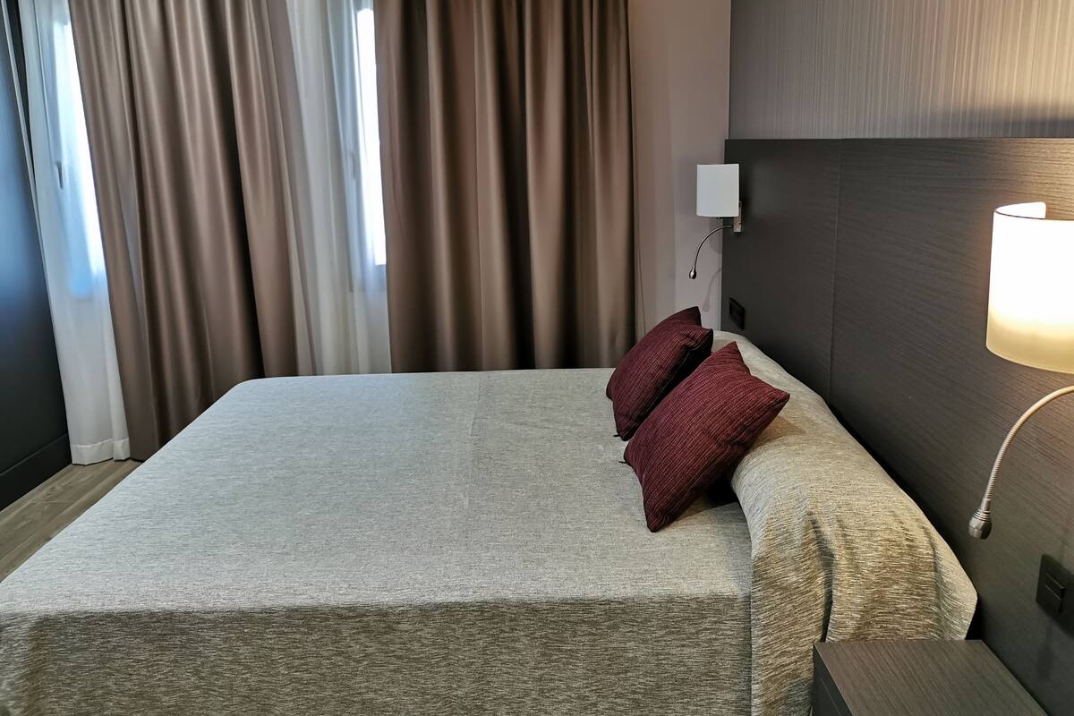 A hotel room with a large bed, a nightstand, a lamp, and a view of the outside through curtains.