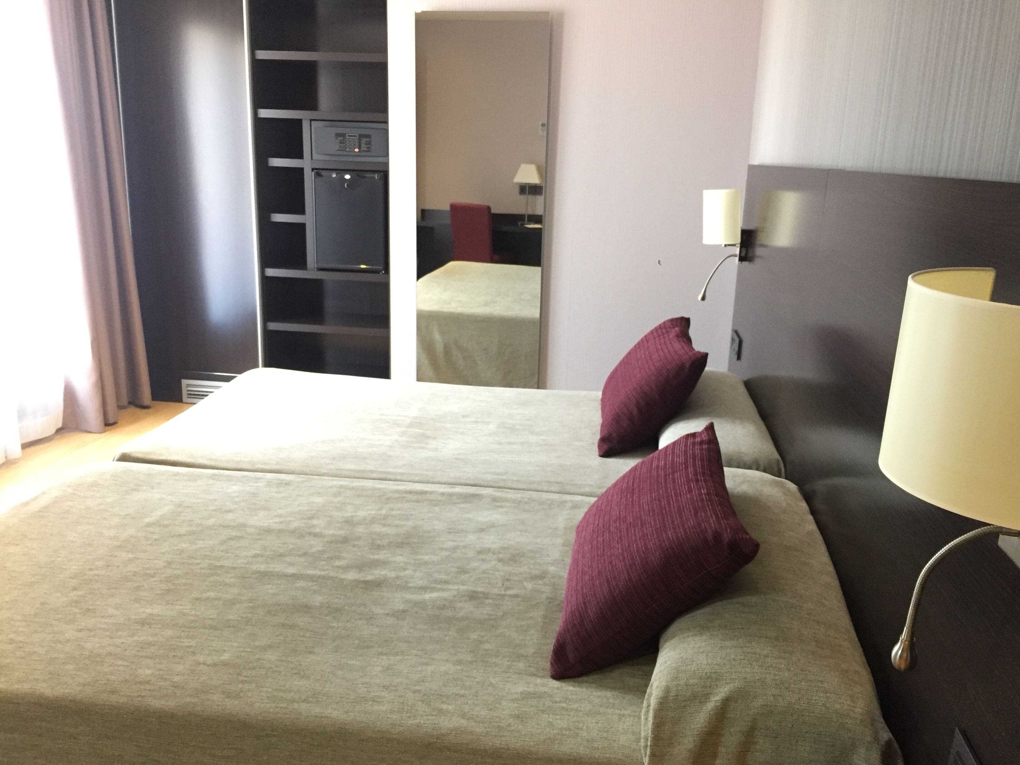 A hotel room with a bed, two pillows, a bedside table, a lamp, a wardrobe, and a mirror.