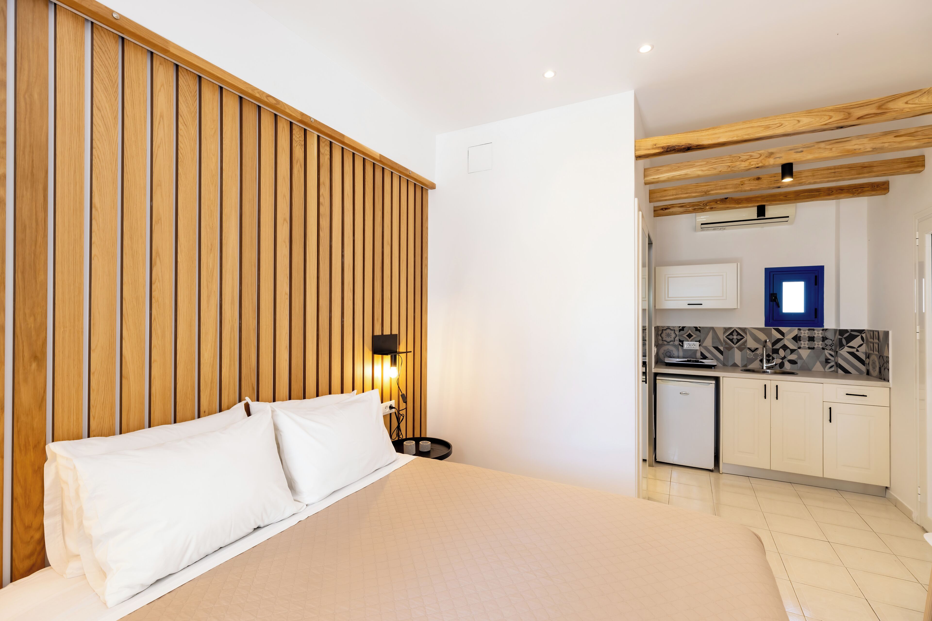 A bedroom with a wooden headboard, a bed with white bedding, a bedside table with a lamp, and a view into a kitchen area with a refrigerator and a microwave.