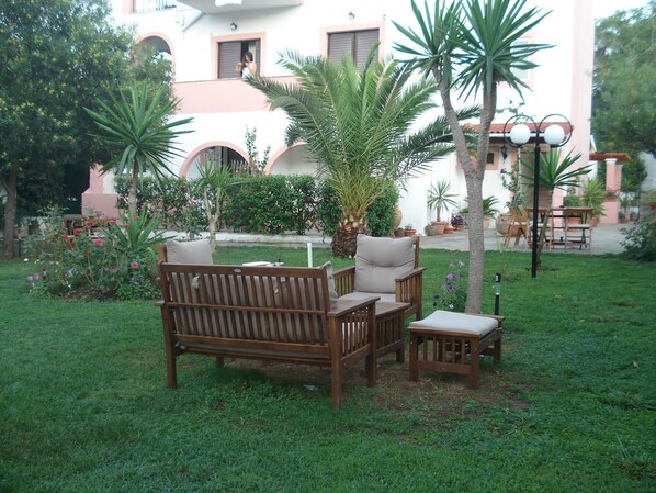 A patio with a wooden bench and table, surrounded by greenery and a building with pink and white walls. - Villa Avra (Corfu)