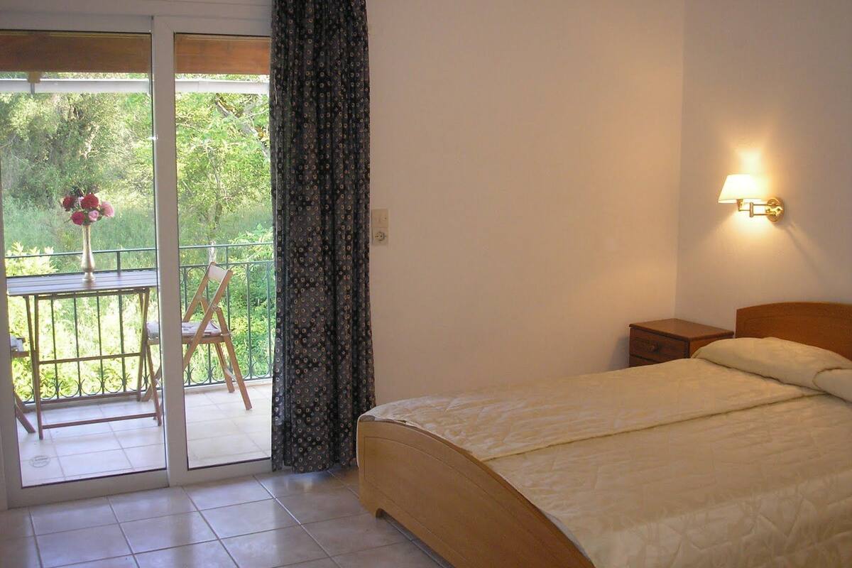 A bedroom with a bed, a nightstand, a lamp, a chair, a table with a vase of flowers, and a view of greenery through a sliding door.