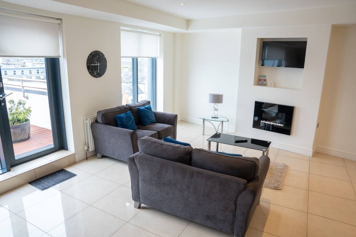 A modern living room with two grey sofas, a glass coffee table, a fireplace, and a large window with a view of buildings.