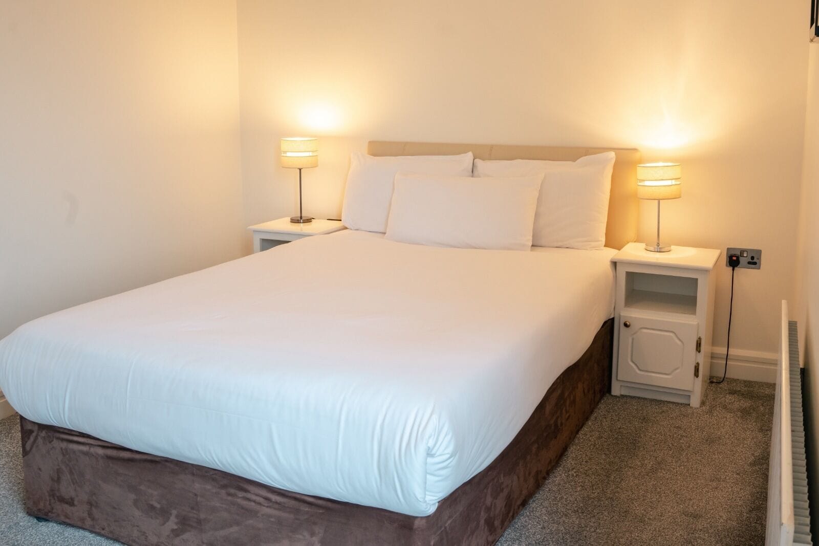 Una cama bien tendida con sábanas blancas, dos mesitas de noche con lámparas y un pequeño mueble bajo la cama.