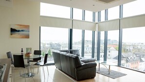 A modern living room with a large window, a grey sofa, a dining table with chairs, and a kitchen area. - Citypoint Apartments (Galway)