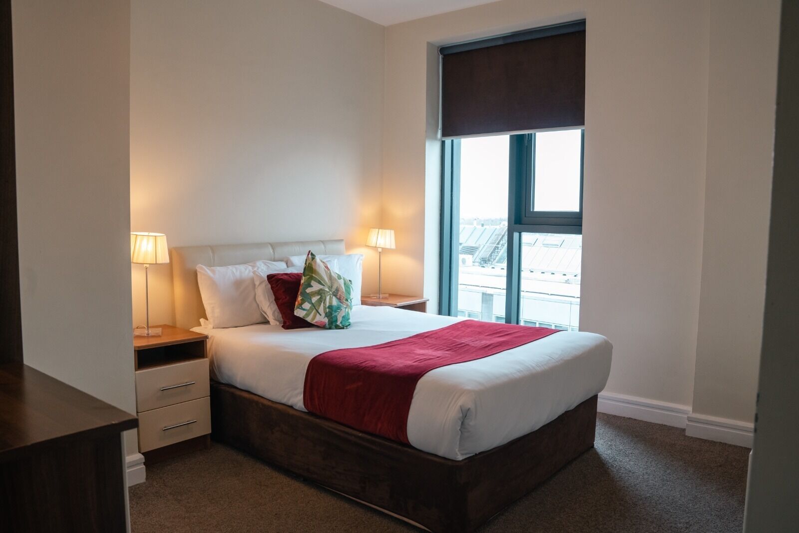 Een net opgemaakt bed met wit en rood bedlinnen, een nachtkastje met een lamp en een raam met uitzicht op gebouwen.