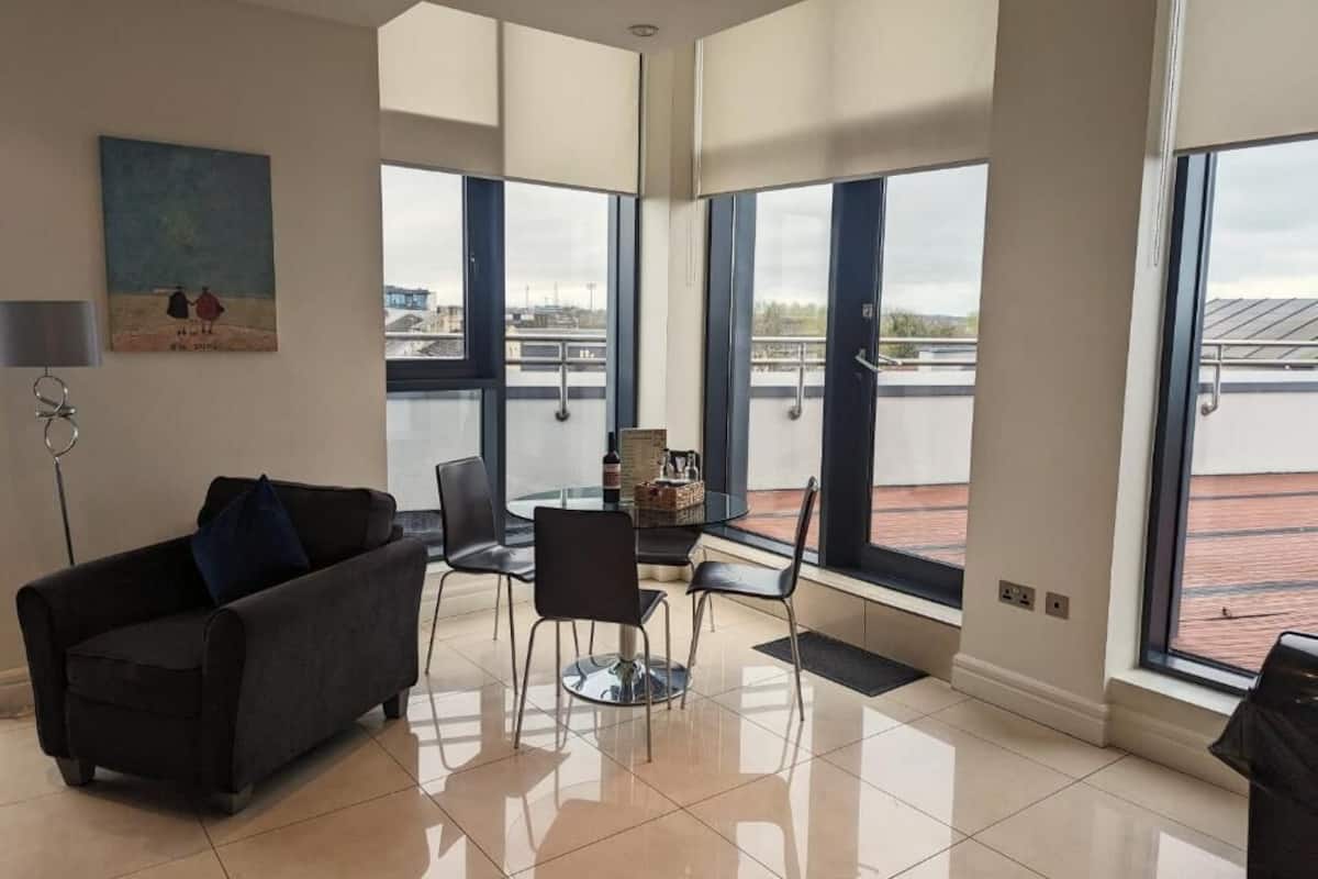A modern living room with a black sofa, a glass coffee table, and a painting on the wall. Large windows offer a view of the cityscape and a bridge.