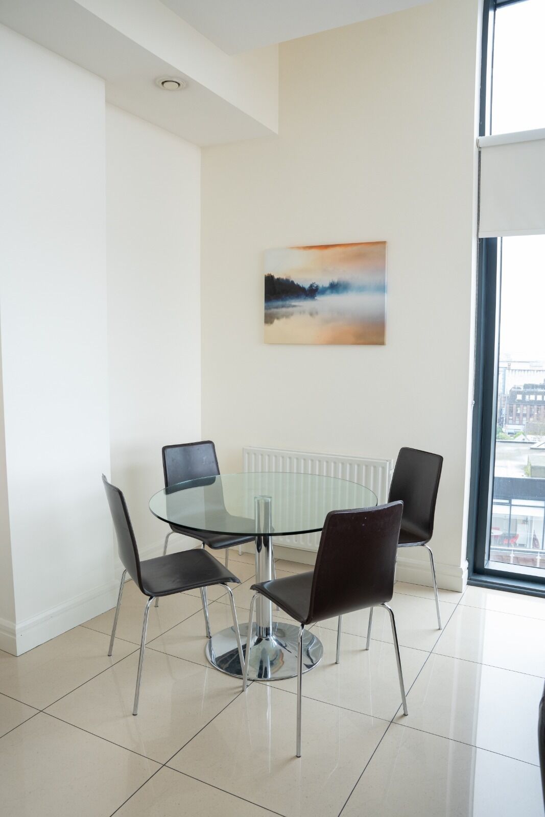 A modern dining area with a glass table and four black chairs. A white radiator is below the table, and a painting hangs on the wall.