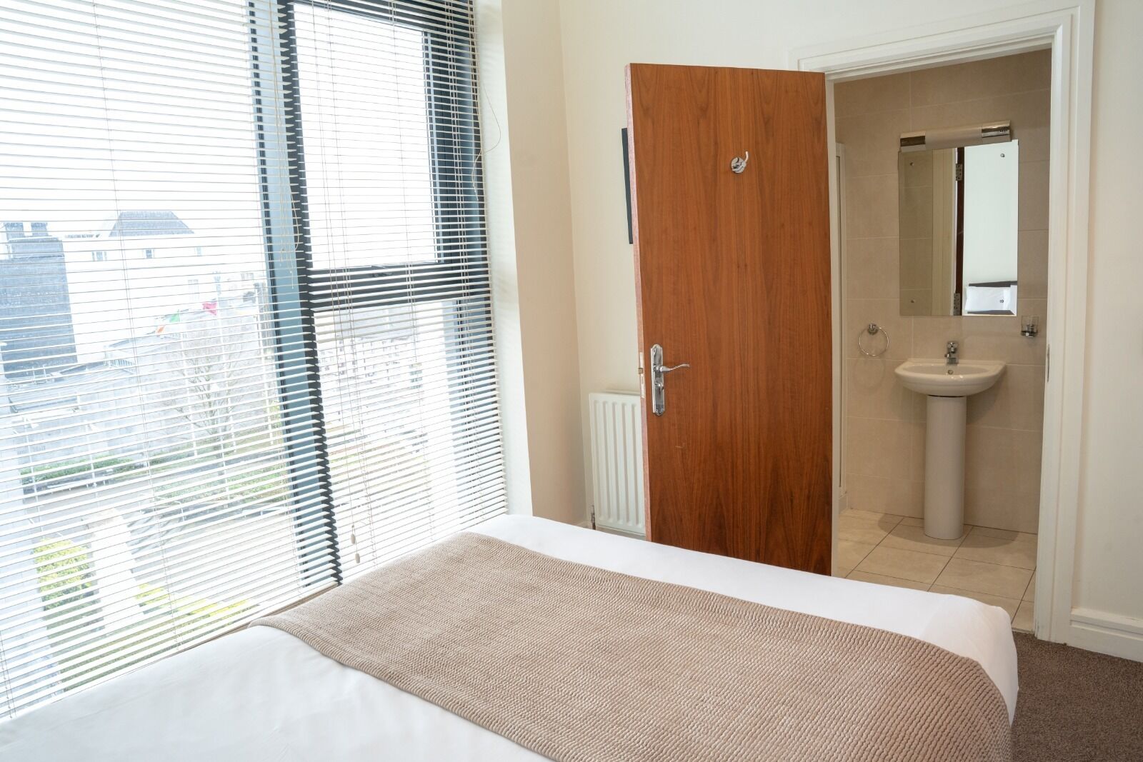 A hotel room with a bed, a wooden door, a bathroom with a sink and mirror, and a window with blinds.