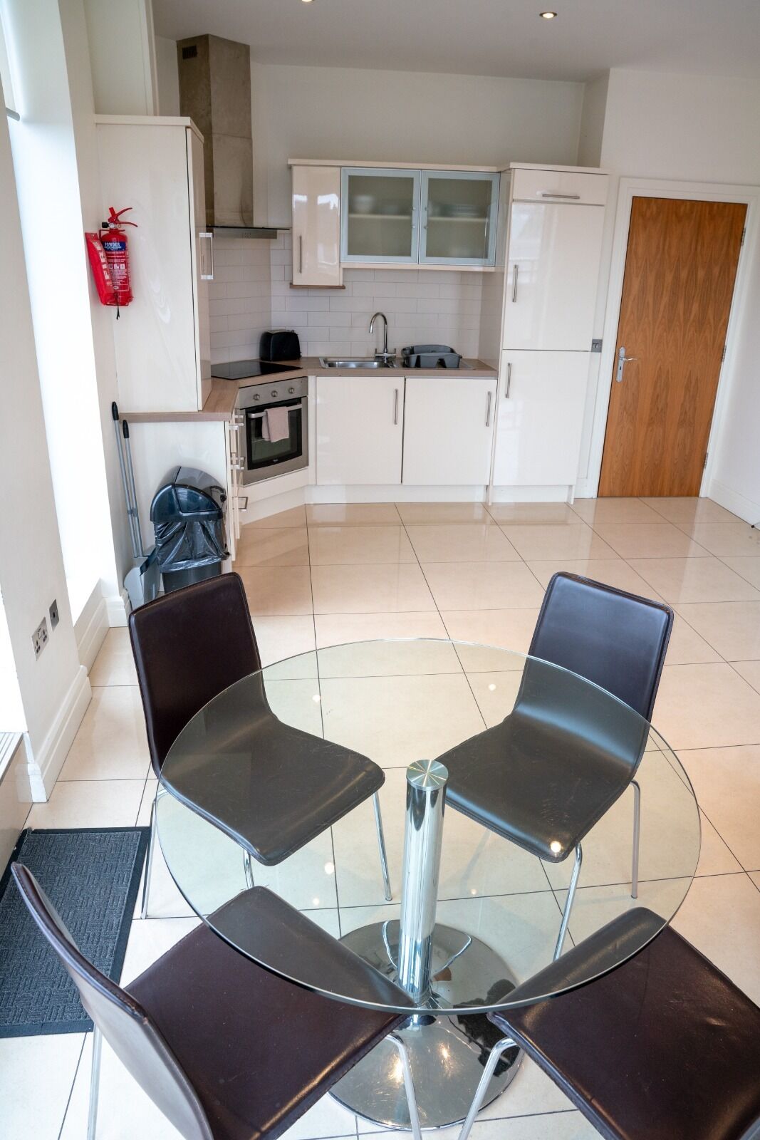 A modern kitchen with white cabinets, a stainless steel sink, and a built-in oven. There is a glass table with four chairs in the foreground.