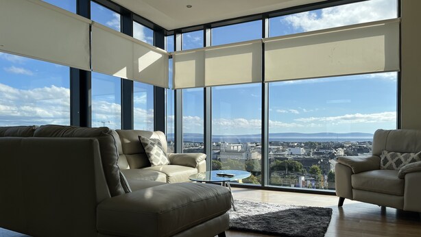 A spacious living room with a large window offering a city view, a beige sofa, a small round table, and a patterned throw pillow. - Citypoint Apartments (Galway)