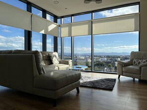 Ein geräumiges Wohnzimmer mit großem Fenster und Blick auf die Stadt, ein beiges Sofa, ein kleiner runder Tisch und ein gemustertes Kissen.
