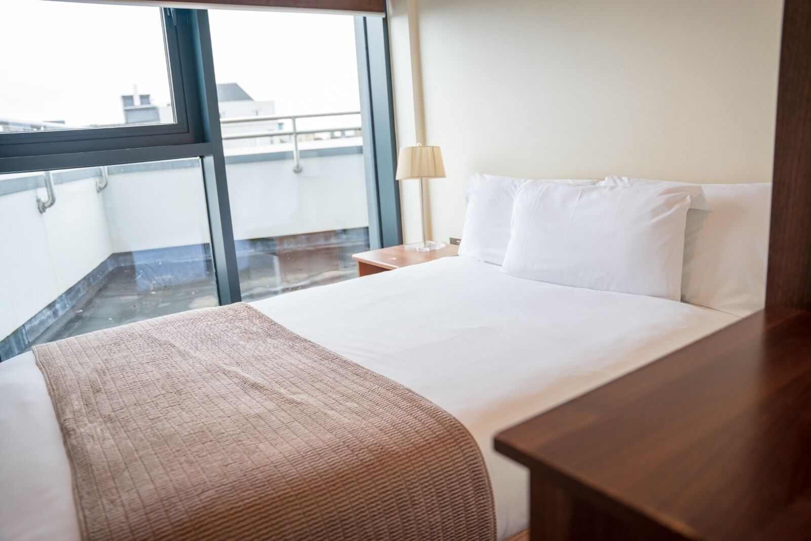 Quarto de hotel com uma cama grande, uma mesa de cabeceira de madeira, uma luminária e vista da cidade através de uma janela ampla.