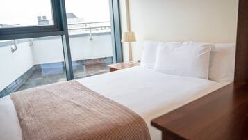 Quarto de hotel com uma cama grande, uma mesa de cabeceira de madeira, uma luminária e vista da cidade através de uma janela ampla.