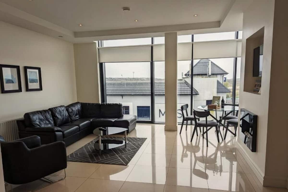 A modern living room with a black leather sofa, a coffee table, and a dining area with chairs. Large windows offer a view of the outside.